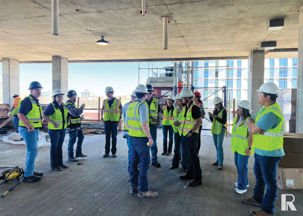 A group of people wearing hard hats and high-visibility vests stand on an unfinished concrete floor at a construction site.