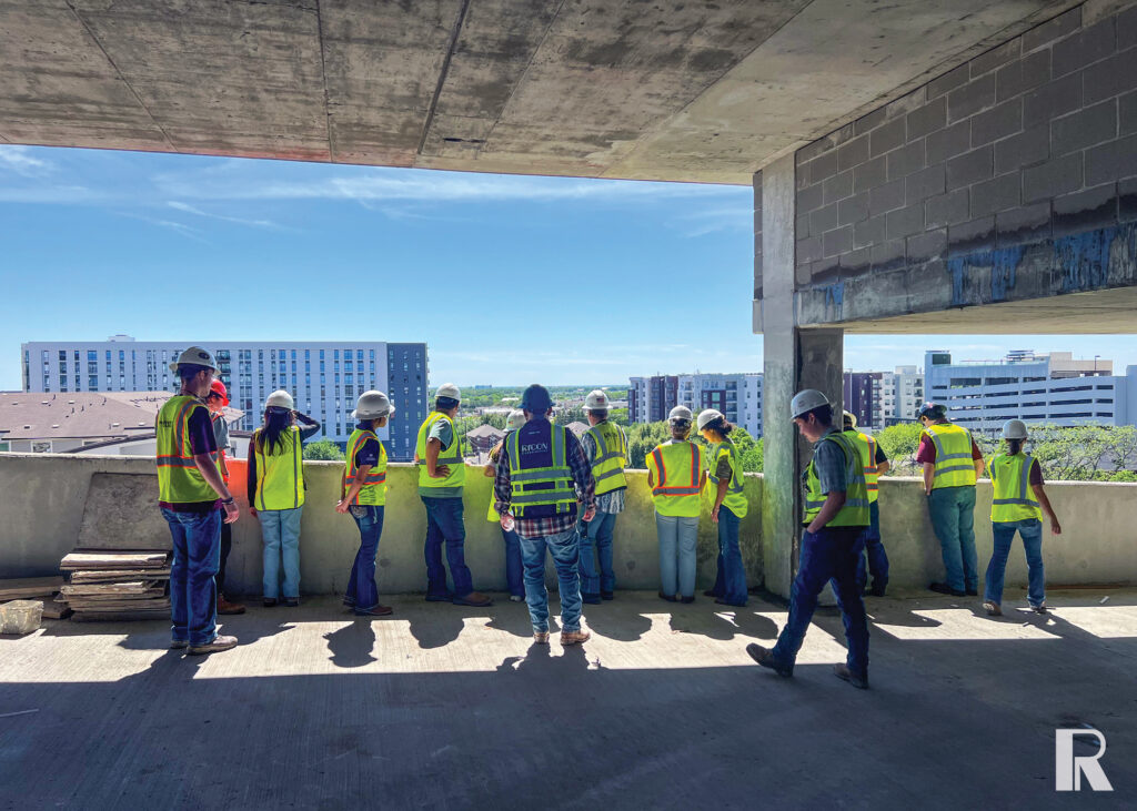 A group of construction workers in hard hats and high-visibility vests stand at the edge of an unfinished concrete building, looking out at a city skyline.