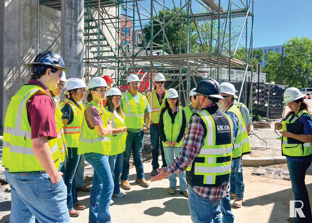 A group of people in hard hats and high-visibility safety vests gather at a construction site while one person speaks to the group.