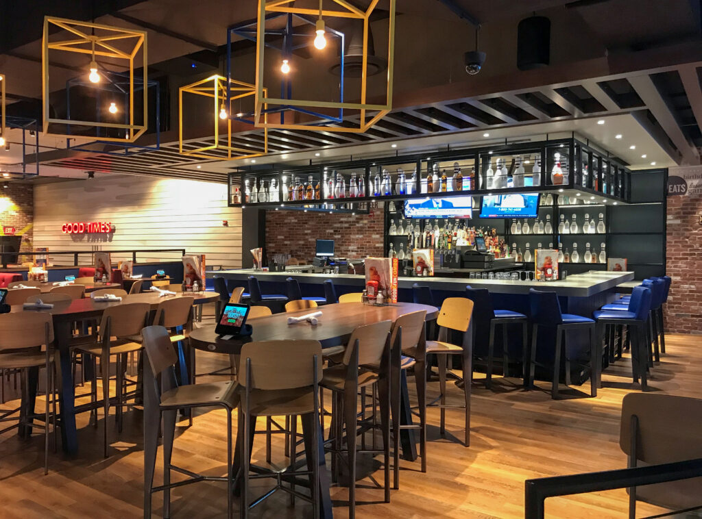 Empty tables and chairs in a restaurant with a bar, wood floors, and geometric light fixtures.