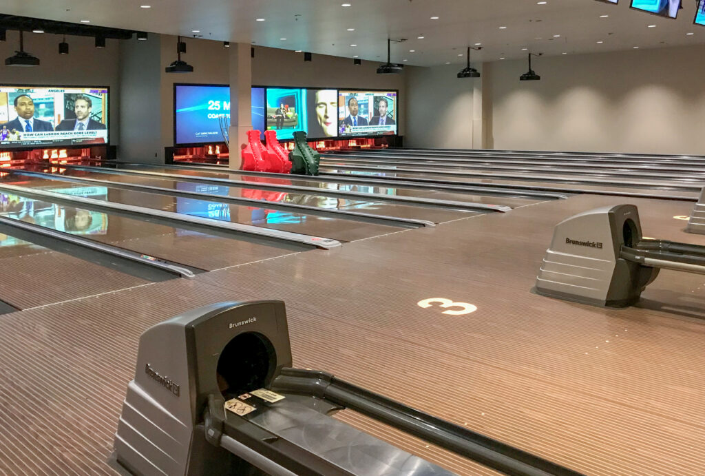 An indoor bowling alley with multiple lanes, ball returns, and large screens displaying sports news.