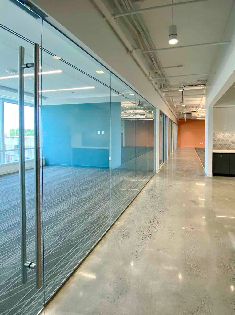 A long hallway with polished concrete floors, lined with glass-walled office suites painted in blue and orange.