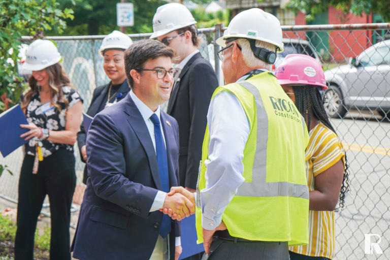 A man in a suit shakes hands with a man wearing a white hard hat and yellow construction vest, while three others stand nearby.