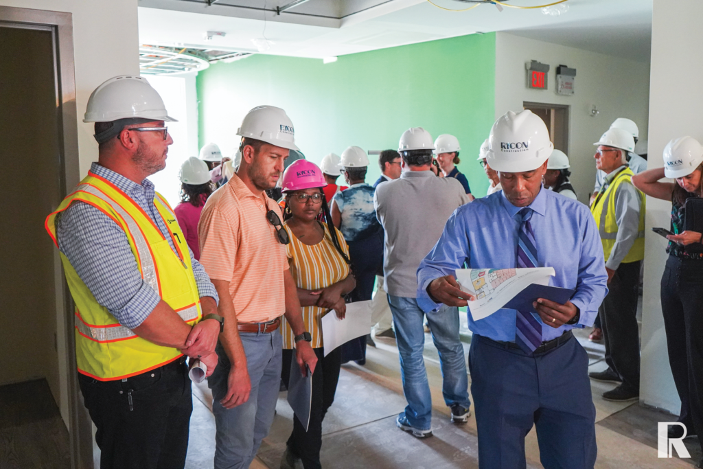 A group of people wearing hard hats and construction gear in an unfinished building, with one man in the foreground reviewing a floor plan document.