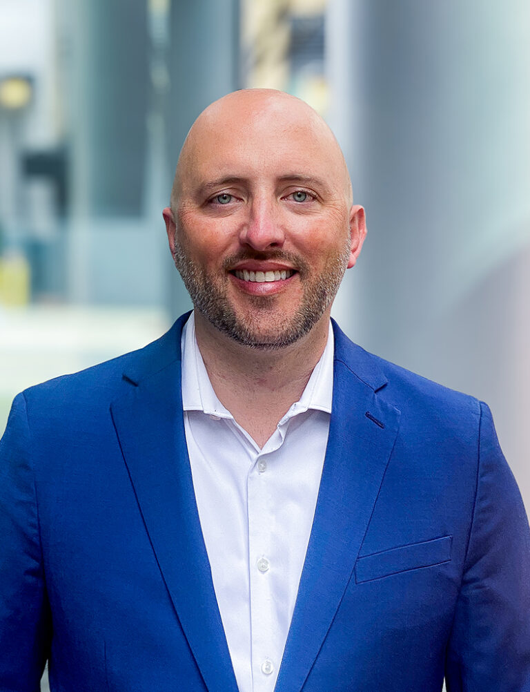 A smiling man with a shaved head and a beard, wearing a blue blazer and a white dress shirt.