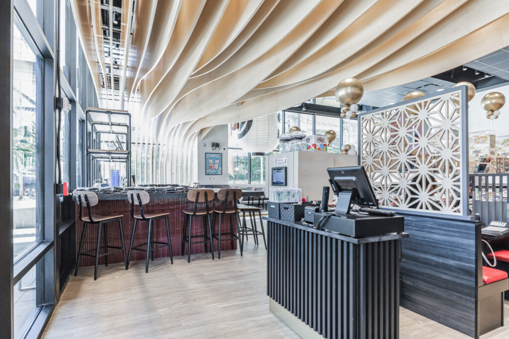 Interior of a restaurant with a curved wooden ceiling, a bar counter with stools, and a checkout station in the foreground.