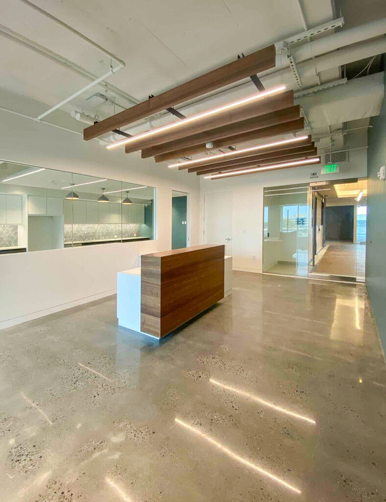 An empty office reception area with a wooden front desk, polished concrete floors, and linear pendant lighting suspended from the ceiling.