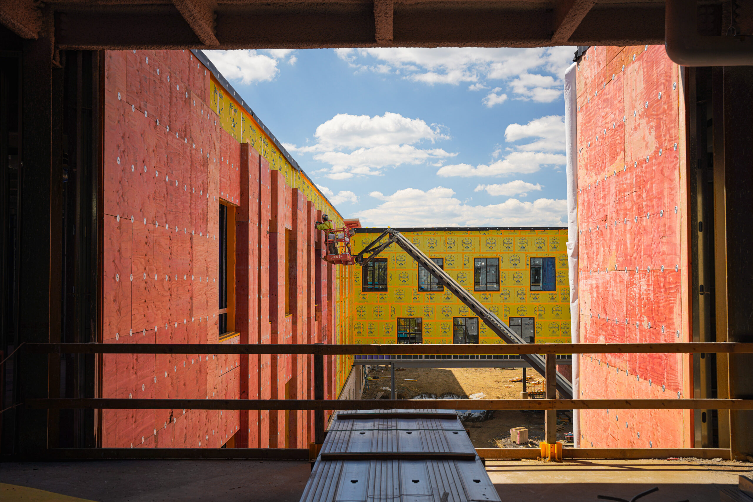 A construction worker in a cherry picker works on the exterior of a building under construction, framed by unfinished interior walls.