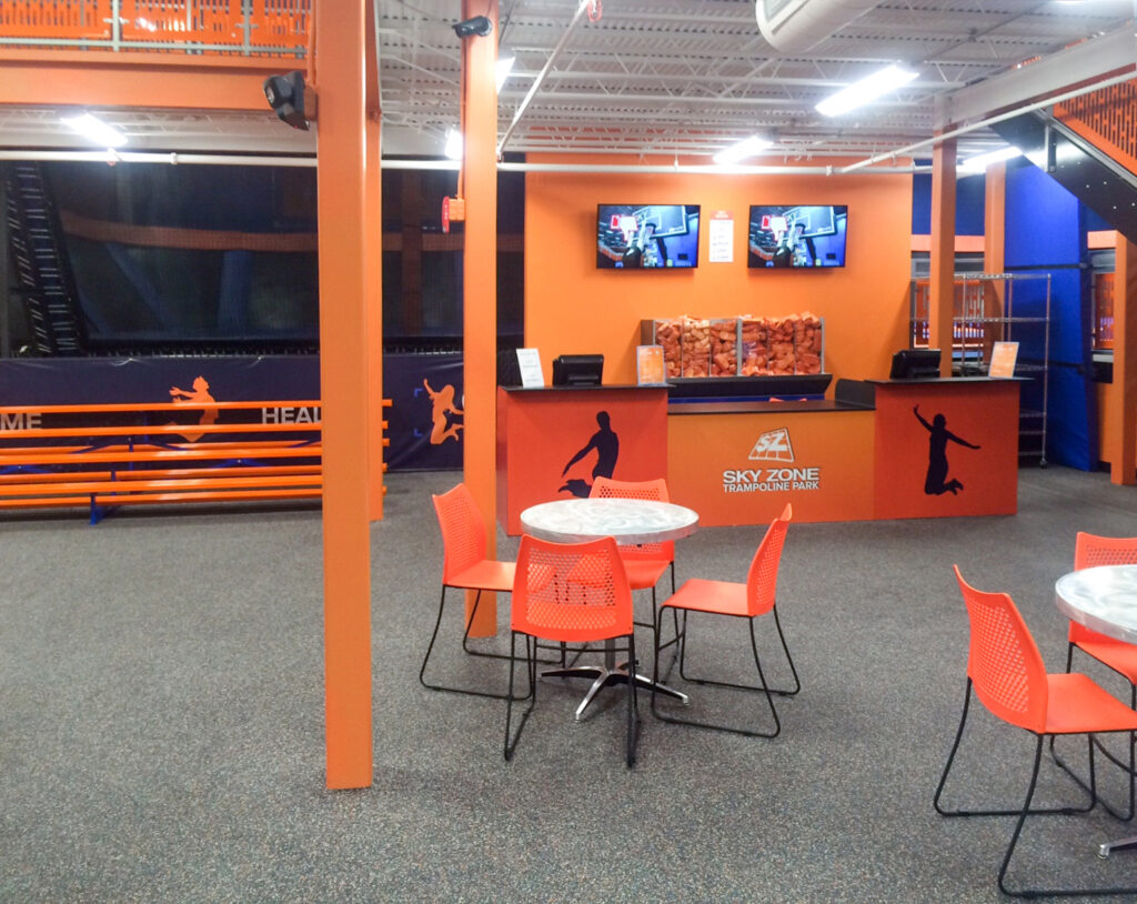 Reception desk for Sky Zone Trampoline Park, featuring orange walls, a seating area with chairs and tables, and two wall-mounted television screens.