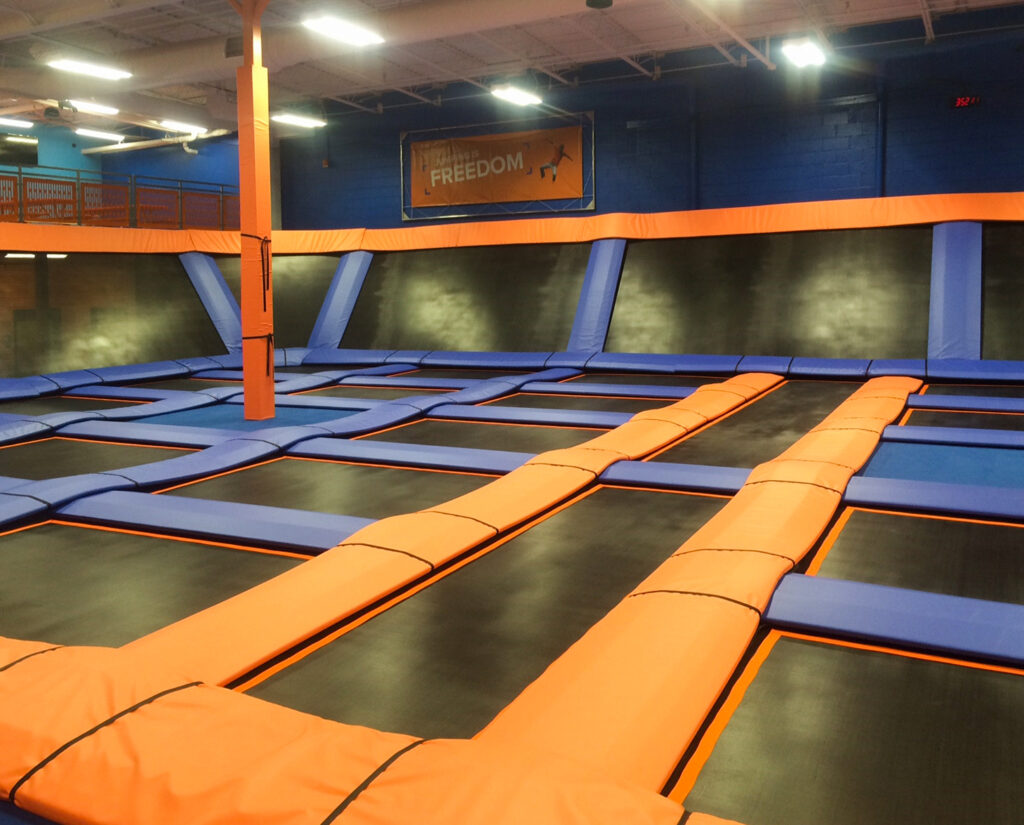 An indoor trampoline park with a grid of black jumping surfaces separated by orange and blue padded frames.