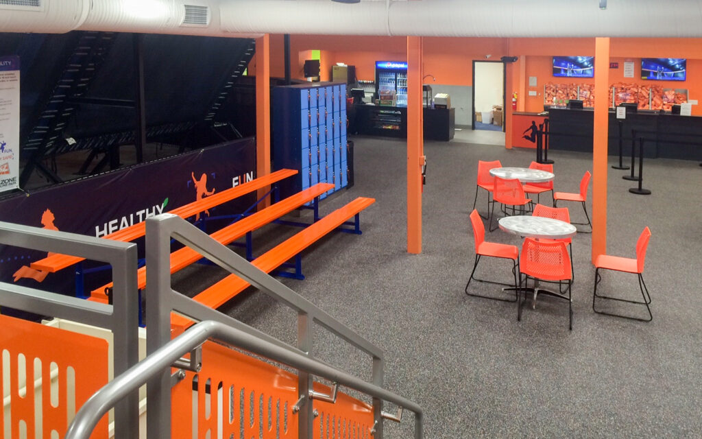 Indoor recreational facility with orange spectator benches, cafe tables with chairs, lockers, and a check-in desk.
