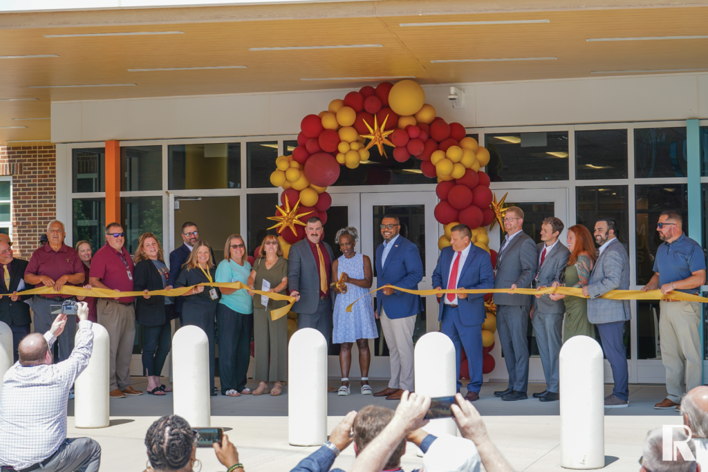 A group of people standing in a line holding a gold ribbon across the entrance of a building decorated with a balloon arch for a ribbon-cutting ceremony.