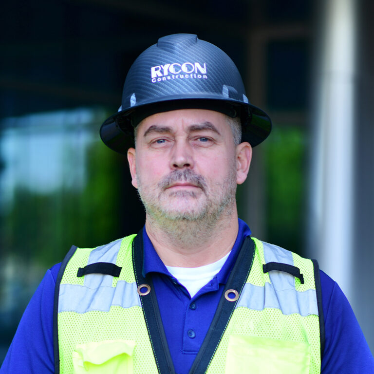 A construction worker wearing a hard hat with the Rycon Construction logo and a yellow high-visibility safety vest.