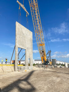A large yellow crane lifting a precast concrete wall panel at a construction site.