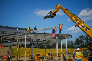 Two construction workers guide a steel beam labeled “STEEL VALLEY ELEMENTARY SCHOOL” into place with an American flag attached, hoisted by a crane.