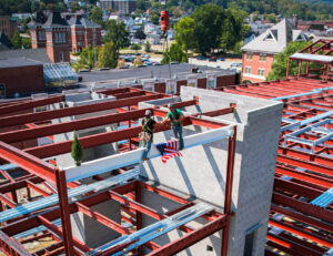 Two construction workers high-five while standing on a steel beam being lifted by a crane, with a small American flag attached to the beam.