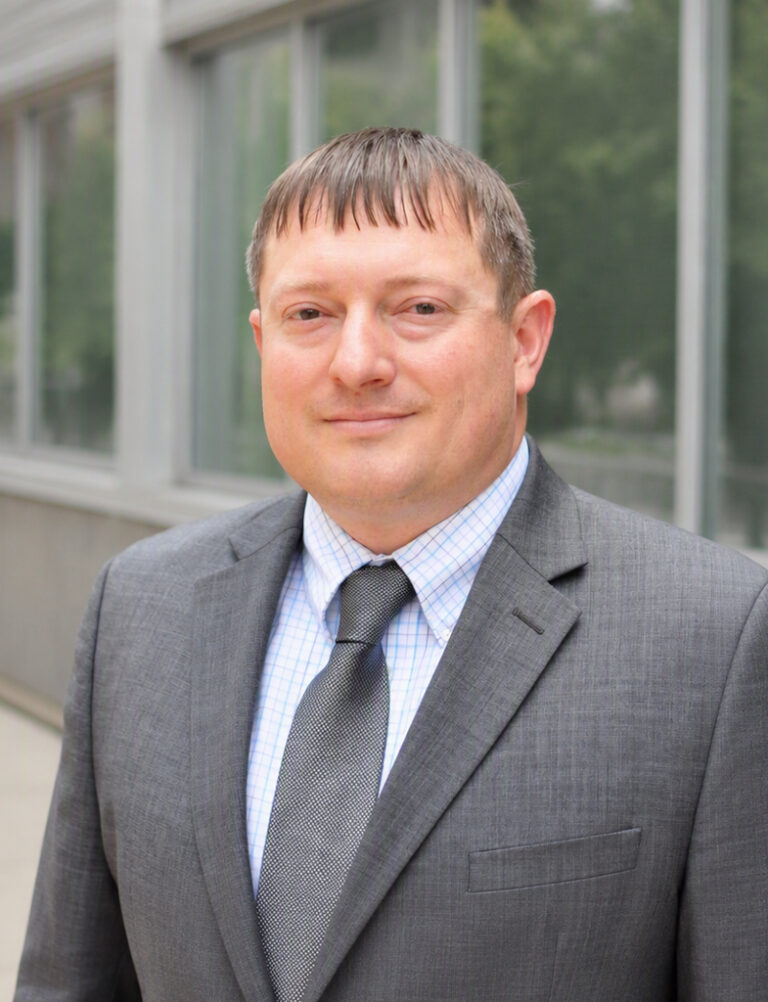 A middle-aged man in a grey suit, light blue checkered shirt, and grey tie.