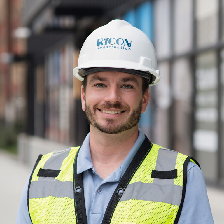 A smiling man wearing a white Rycon Construction hard hat and a high-visibility safety vest.