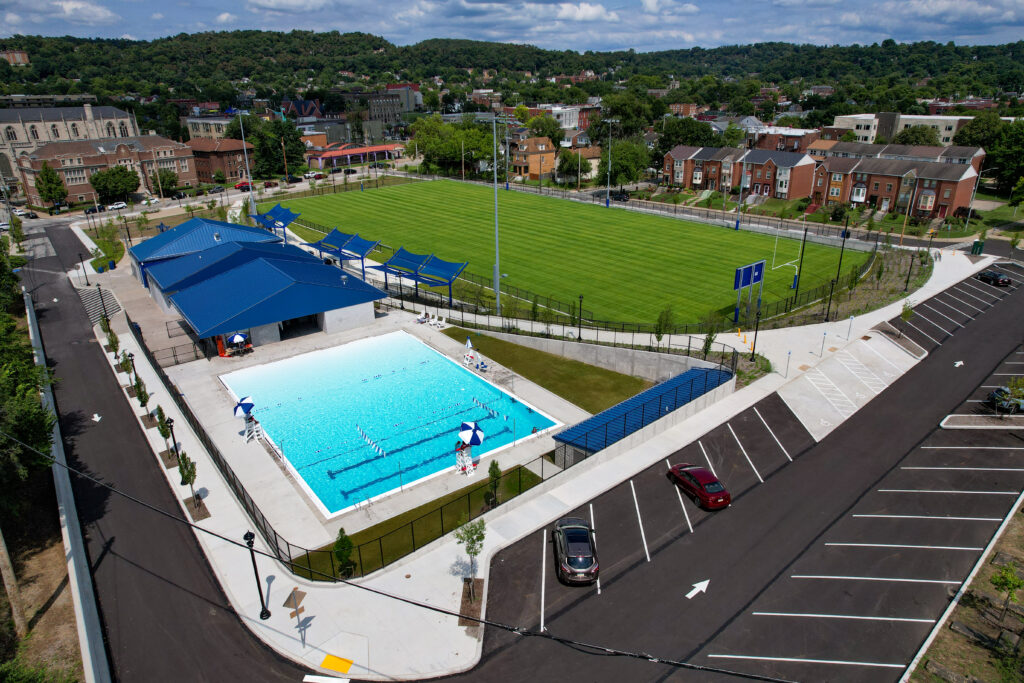Aerial view of an outdoor swimming pool and a green athletic field next to a parking lot in a residential neighborhood.