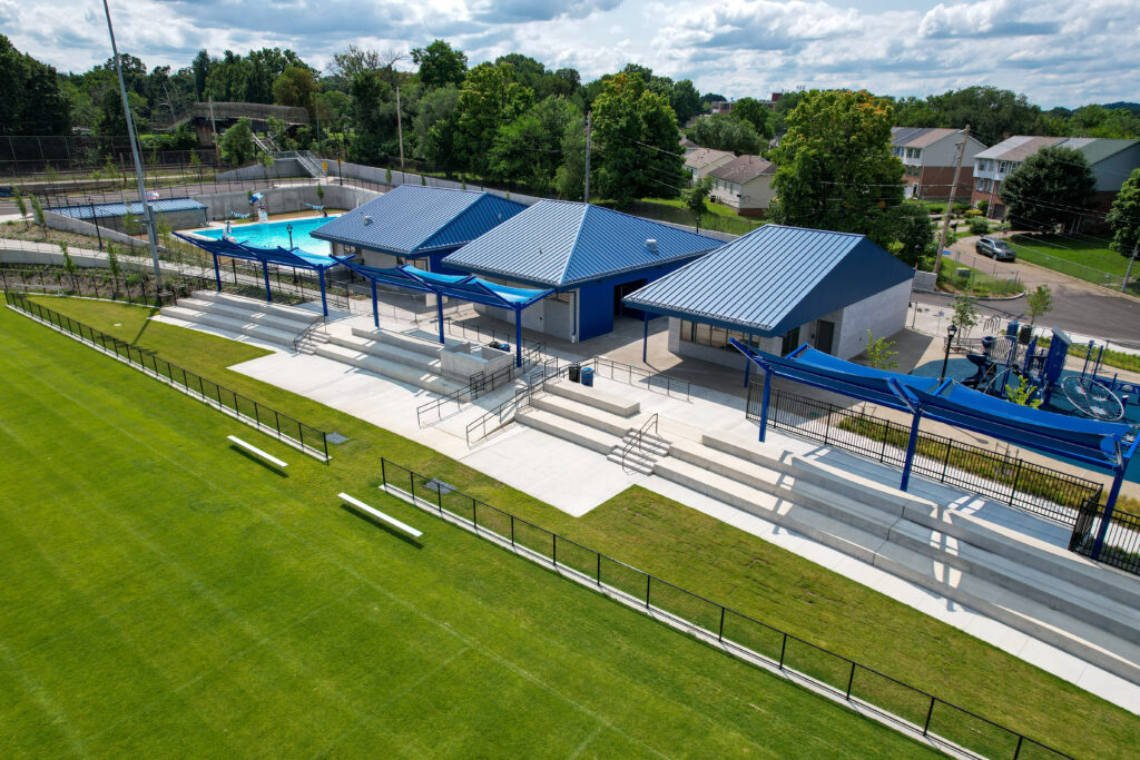 An outdoor recreation area with a swimming pool, three blue-roofed buildings, and concrete tiered seating overlooking a large grass field.
