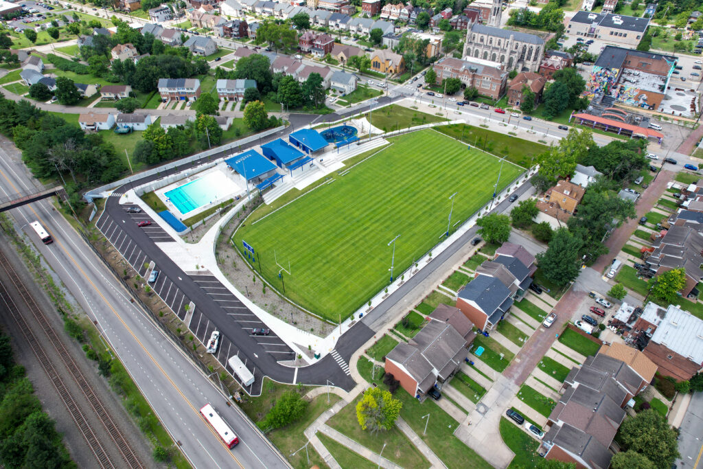 Aerial view of a public park featuring a rectangular sports field, a swimming pool with a blue canopy, and an adjacent parking lot in a residential neighborhood.