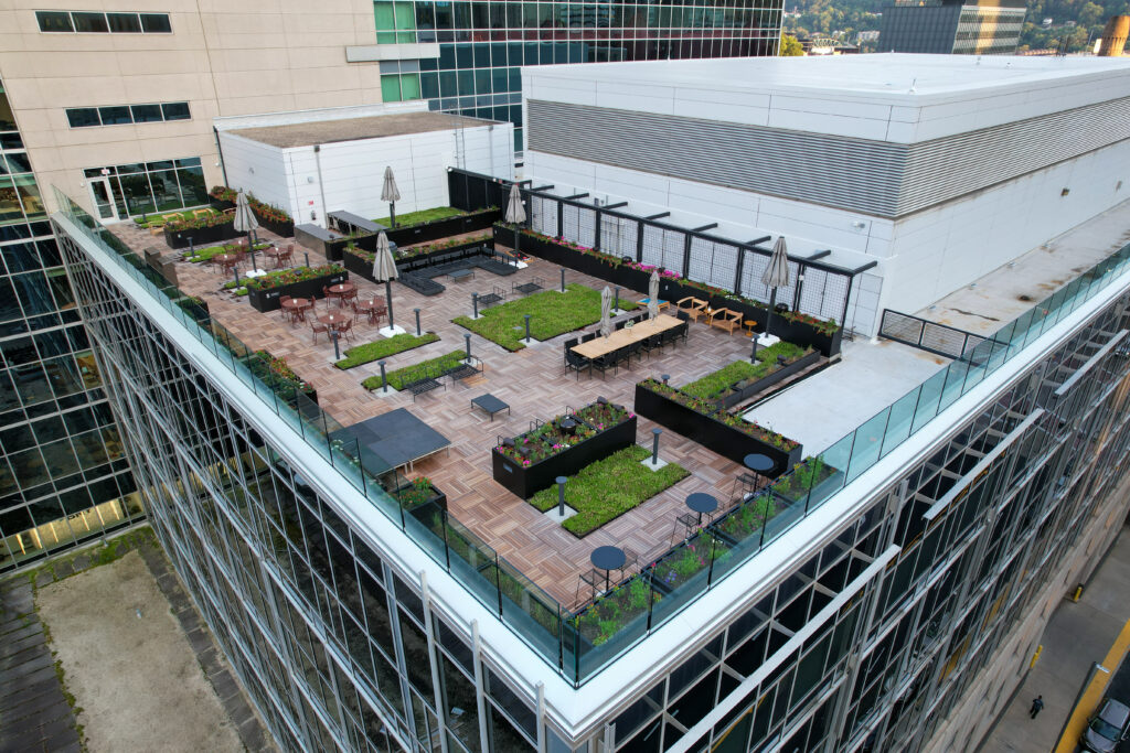Aerial view of a rooftop terrace featuring wooden decking, planters with flowers, dining tables with umbrellas, and a ping pong table, enclosed by glass railings.