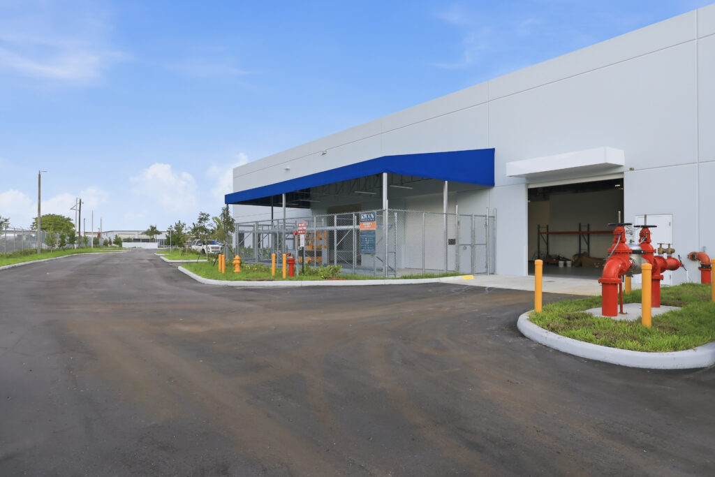 A modern industrial warehouse building featuring a fenced service entrance with a blue awning and a red fire department connection in the foreground.