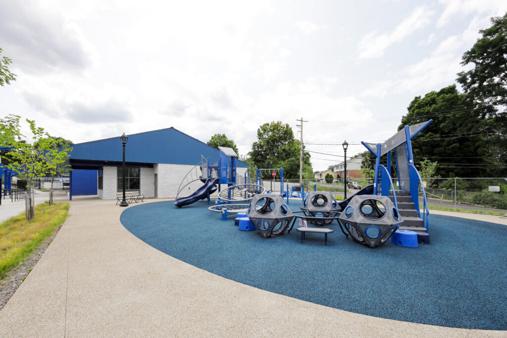 Outdoor playground with blue rubber flooring, climbing structures, and a slide next to a blue and white building.