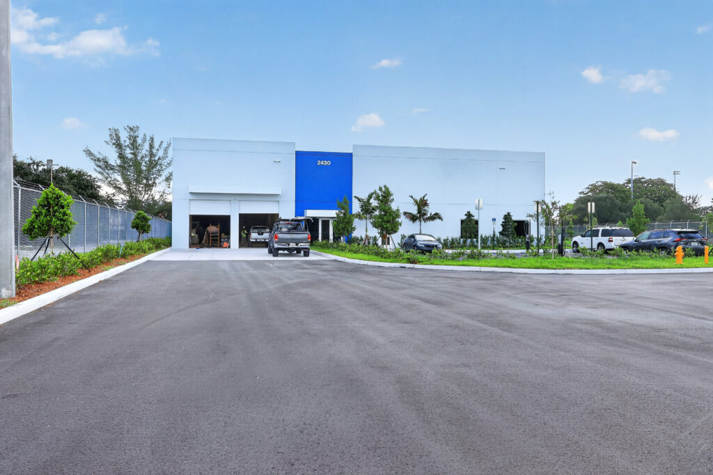 A white and blue commercial warehouse building with the address 2430, fronted by a parking lot with several vehicles.