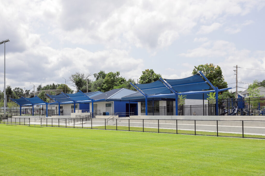 A field house with blue shade structures and a black metal fence, bordering a green grass field.