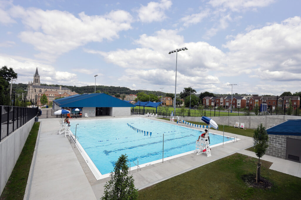 An outdoor swimming pool with lanes, surrounded by a concrete deck, lifeguard chairs, and a pool house under a cloudy sky.