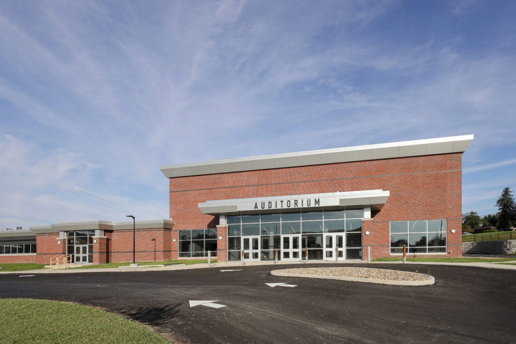 Exterior of a brick school auditorium with a glass entrance, facing a paved parking lot.
