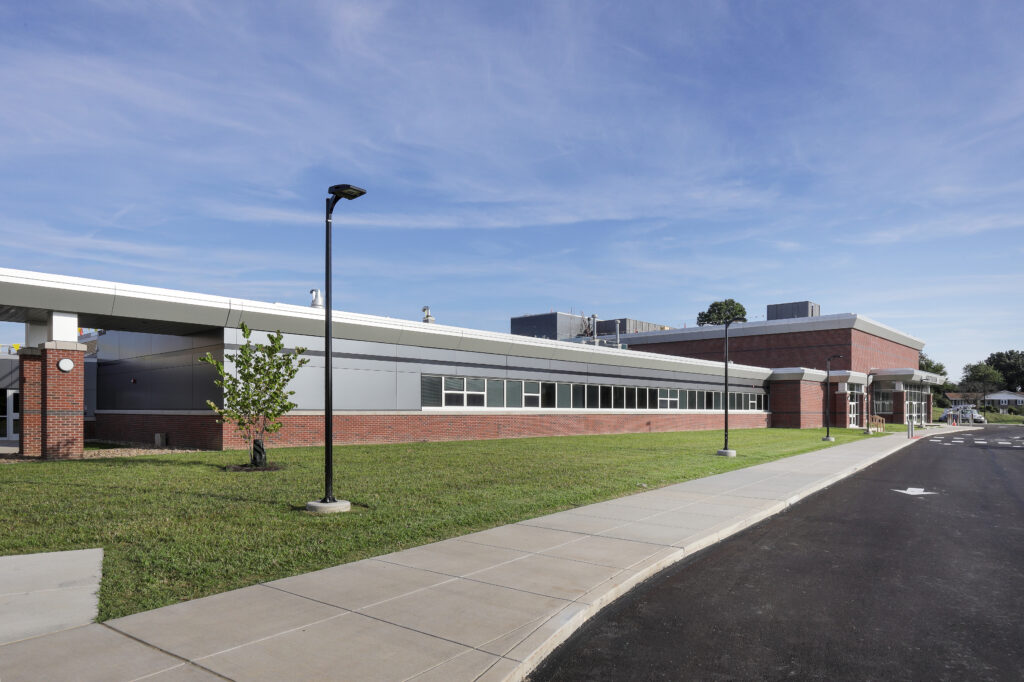 Exterior of a single-story brick and metal school building with a front sidewalk and parking lot.