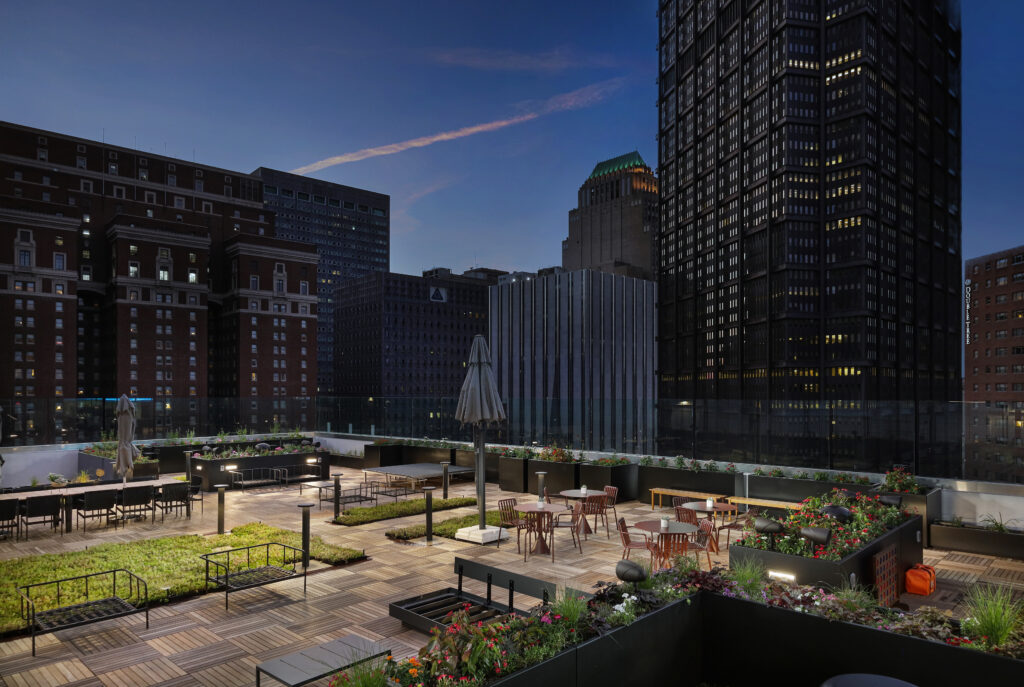 Rooftop terrace with wooden decking, dining tables, and planters, surrounded by city skyscrapers at dusk.