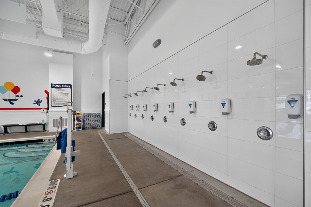 A row of wall-mounted showers and soap dispensers along a tiled wall at a pool deck.