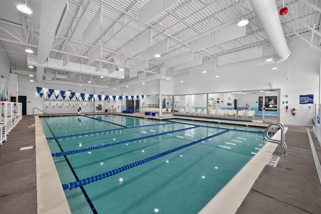 An indoor lap pool with lane dividers, surrounded by a white deck, large windows looking into an adjacent room, and hanging acoustic ceiling panels.
