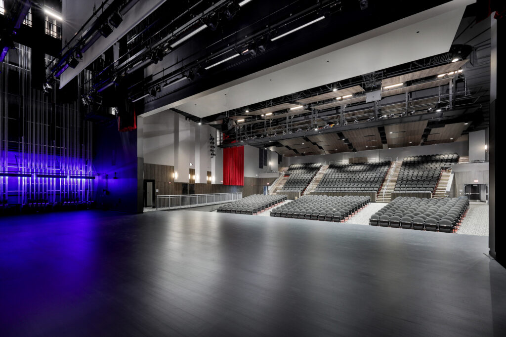 View of an empty theater stage looking out toward tiered seating, with blue accent lighting on the left and stage rigging overhead.