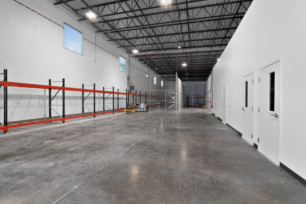 An empty warehouse interior featuring industrial shelving on the left, a fenced enclosure in the back, and a row of white doors on the right.