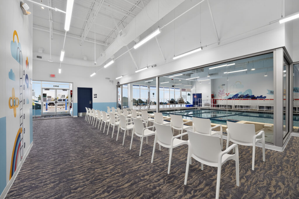 A row of white chairs faces a glass partition looking into an indoor swimming pool.