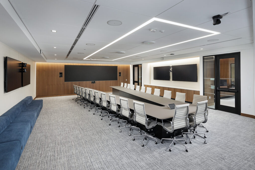 A modern conference room with a long table surrounded by many white ergonomic chairs, wooden wall panels, and large wall-mounted screens.
