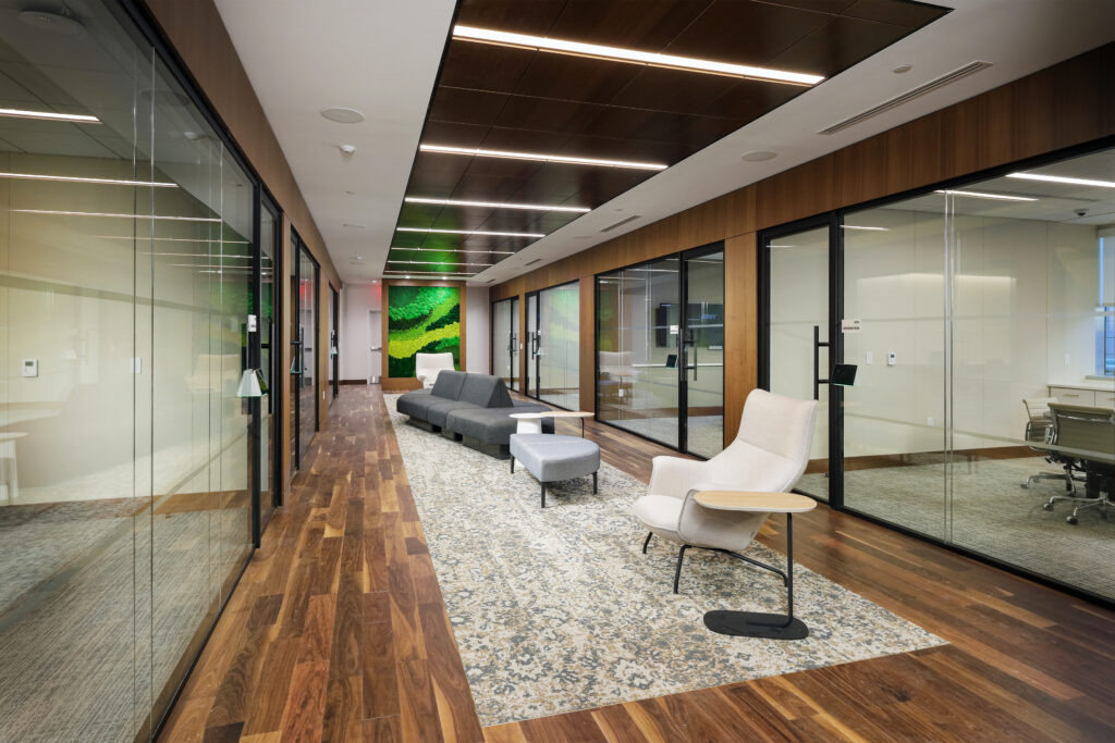 Modern office hallway with glass-walled rooms, a patterned area rug, a grey sectional sofa, and a cream-colored lounge chair with an attached side table.