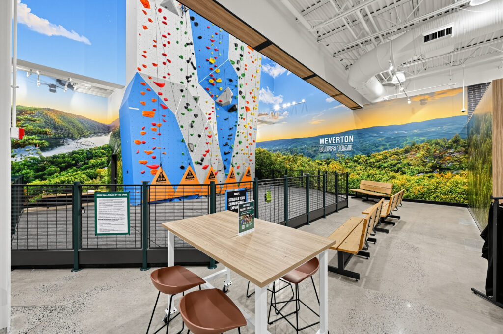 Indoor climbing wall with colorful holds, set against a mural of Weverton Cliffs, with a table and bench seating in the foreground.