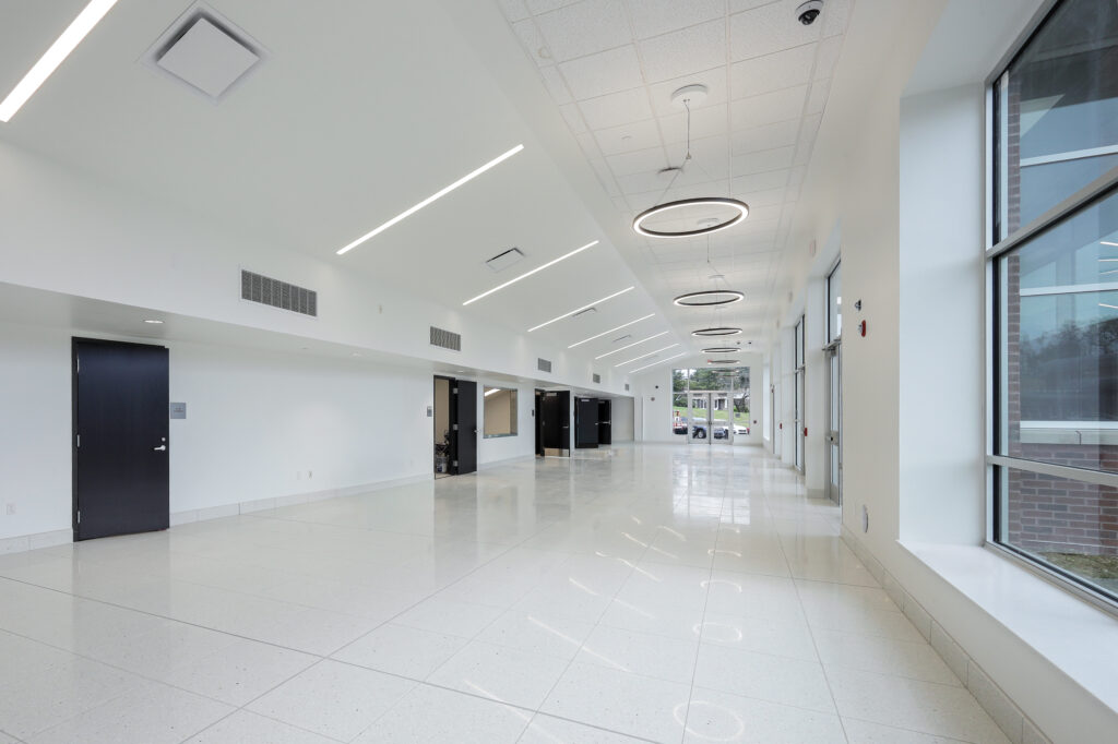 A long, empty white hallway with glossy floors, modern circular pendant lights, and large windows on one side.