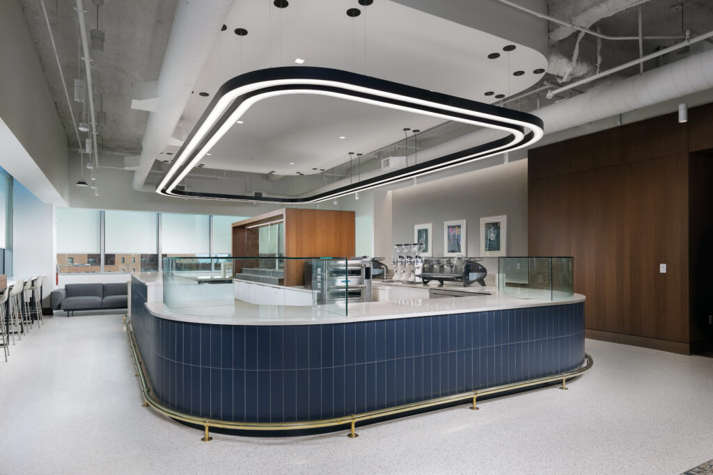 Curved coffee bar with dark blue tiled base and white countertop, featuring an overhead custom light fixture and espresso equipment.