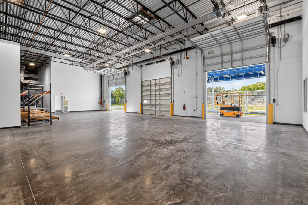Empty industrial warehouse space with concrete floors, high ceilings, and open roll-up doors showing an orange scissor lift outside.