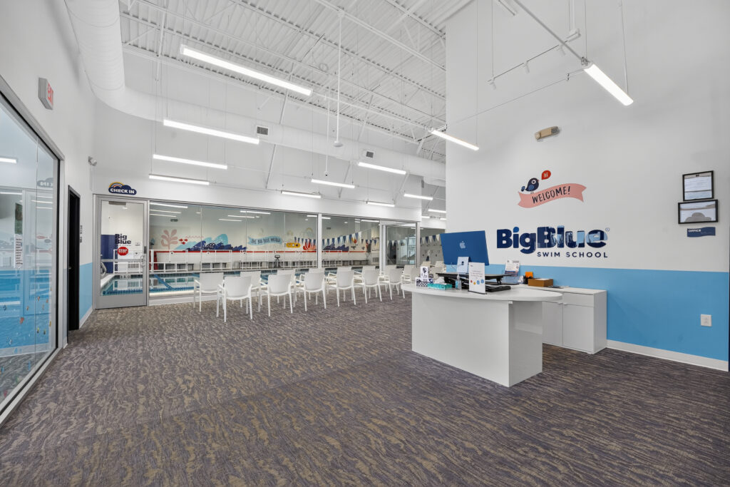 Reception area for Big Blue Swim School with a white desk, waiting chairs, and a view into the indoor pool through large windows.