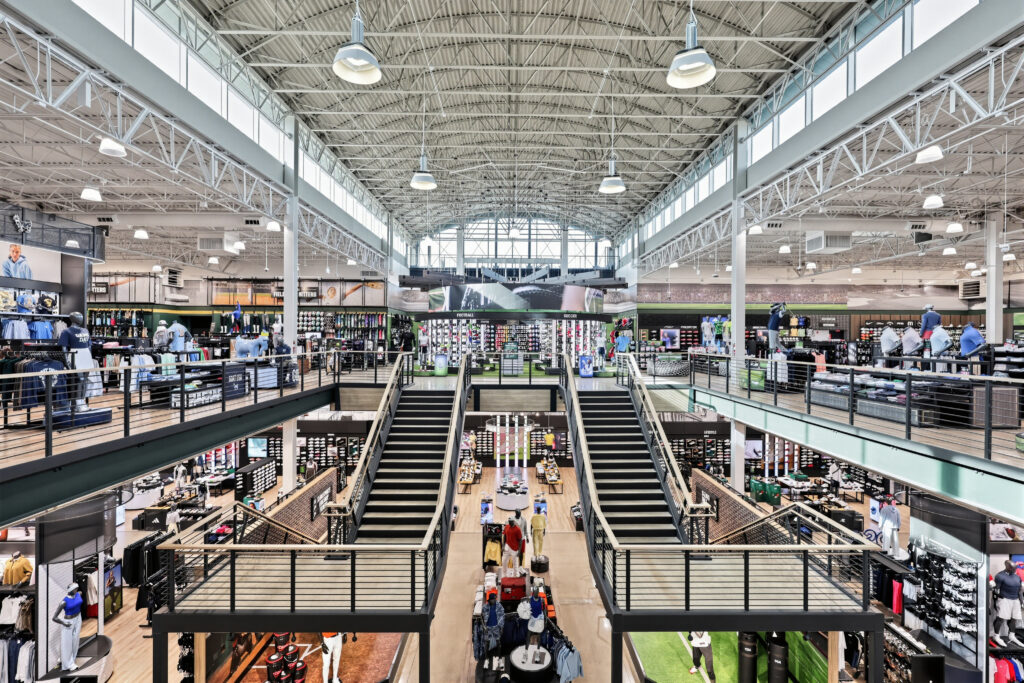 Interior of a two-story sporting goods store featuring a central staircase, clothing displays, and merchandise aisles.