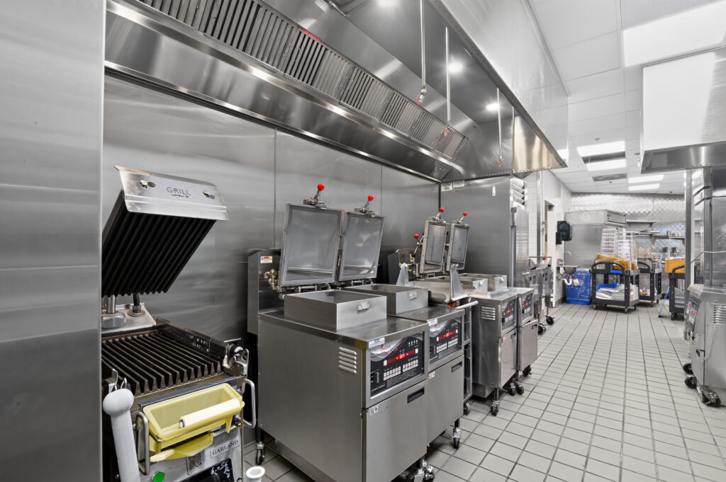 Commercial kitchen with a grill and multiple deep fryers positioned under a stainless steel vent hood.