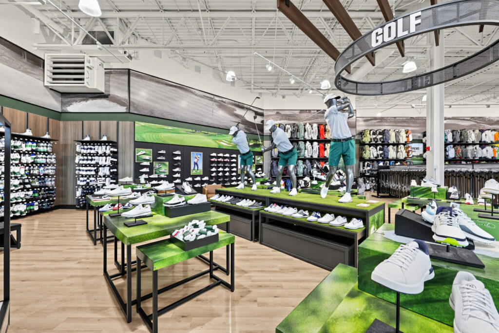 A golf retail store displaying rows of athletic shoes on green turf-covered tables, with mannequins in golf attire posed mid-swing.