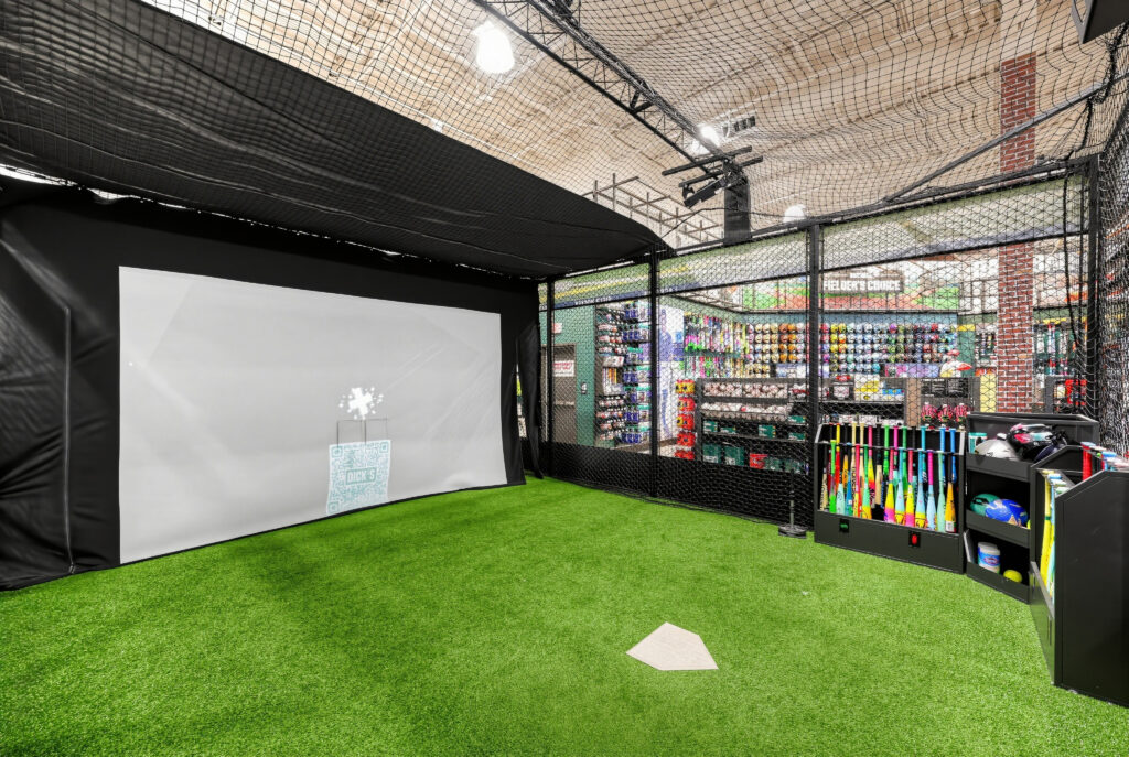 Indoor batting cage with a large projector screen, turf floor, home plate, and a rack of baseball bats.